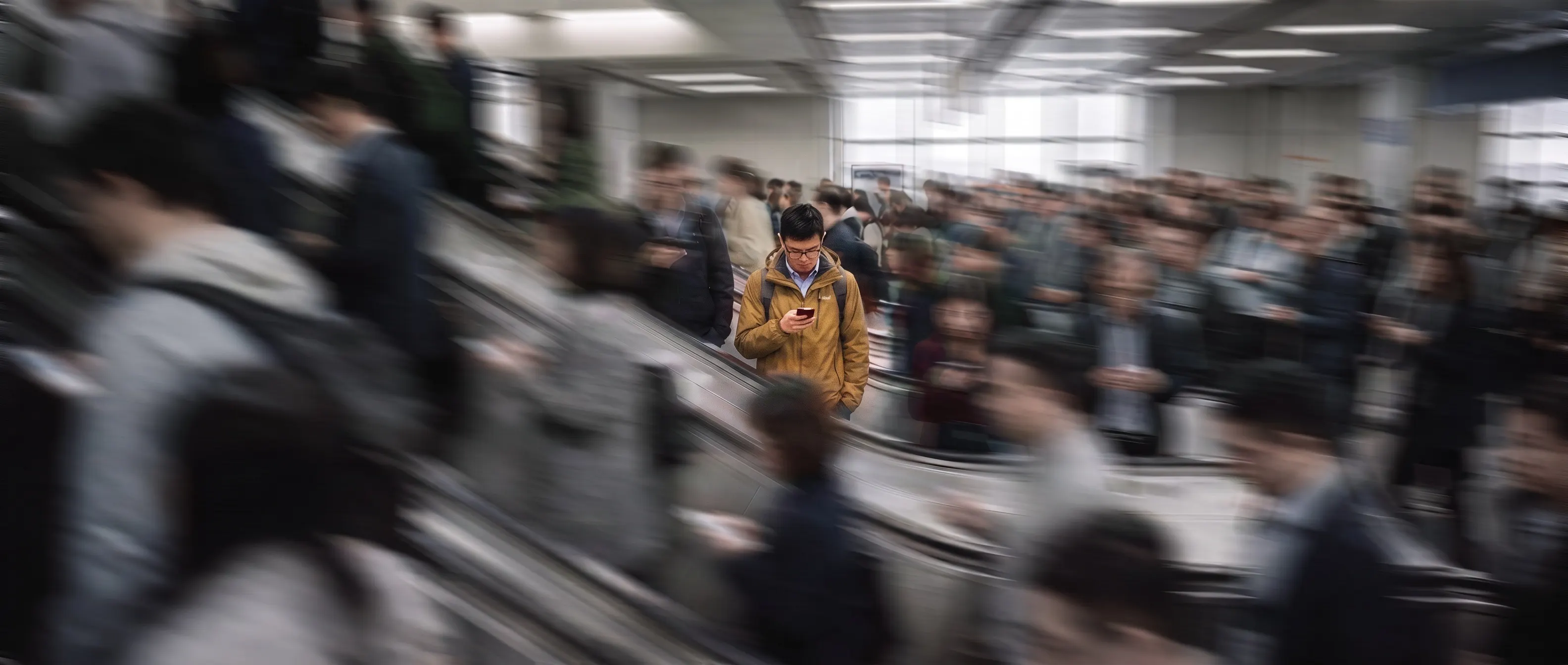 Rolltreppen im Pendlerstrom, alles verwischt — ein Mann in senfgelber Jacke steht scharf am Smartphone